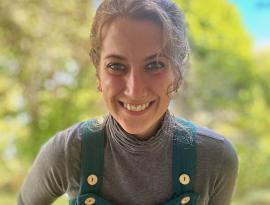 A photo of B Lauer, Executive Director of KLT. She is a white woman with brown hair pulled back into a bun, wearing teal overalls. She is smiling with greenery in the background.